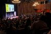 Three people speak on stage while sitting in chairs to a packed auditorium.