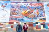 Two people stand in front of a colorful mural. 