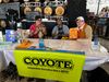 Three people and a dog sit behind a table with a big green banner that says "COYOTE Independent journalism with a bite!" in front of beer kegs
