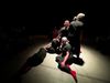 a group of people in black outfits with red socks are positioned on the floor of a grey stage in a spotlight