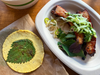 A masa tortilla stamped with a perilla leaf next to a compostable plate with two pork ribs garnished with herbs and slivered cucumber.