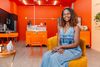 A woman wearing a denim one-piece sits on a yellow upholstered chair in a bright cafe space with orange painted walls.