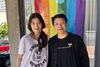 Two women, one with a long ponytail and one with short hair, standing in front of a building with a pride flag hanging from a window.