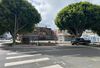 An empty lot on an urban corner is fenced off. Several large trees line its edges. 
