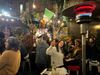 Two women in sweaters hold up peace signs while sitting in a busy bar back patio.
