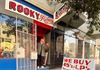 an older white man stands on the porch of a shop with a red and white sign that reads 'Rooky Ricardo's Records.' he is wearing a gray coat and black pants and waving