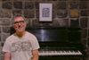 a man in his 50s/60s sits at a piano bench smiling in front of a piano and brick wall