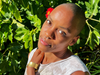 A radiant Black person with a shaved head and a septum piercing standing in the sun against a verdant background. A red flower is tucked behind their right ear.