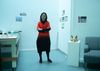 an Asian American woman in a cool red and black dress stands in a blue lit office with a coyote head sculpture on a shelf