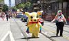Person in a blow-up suit shaped like Pikachu wearing a pride flag marches down a main street next to a person in a white tank top and rainbow hair decor.