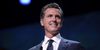 Gavin Newsom, a smarmy looking white man and the governor of California, smiles in a dark blue suit