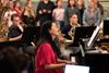 Woman in red shirt playing piano in front of brass instrument players and a group of standing people