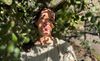 a woman with black hair in a light green shirt and her eyes closed stands with her face dappled in sun and shade under a large plant