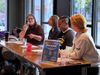 Four women sitting at a wooden table, presenting on a panel. The leftmost woman is speaking into a microphone.