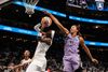 Basketball player in white jersey holding a ball up toward a hoop while another player in a lavender jersey reaches up to block the shot.