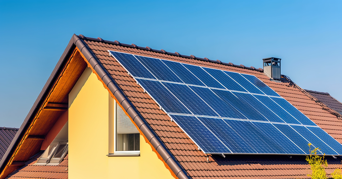 Imagem de uma casa com telhado de telhas cerâmicas marrons e painéis solares instalados. O céu azul sugere um dia ensolarado, destacando a geração de energia sustentável.