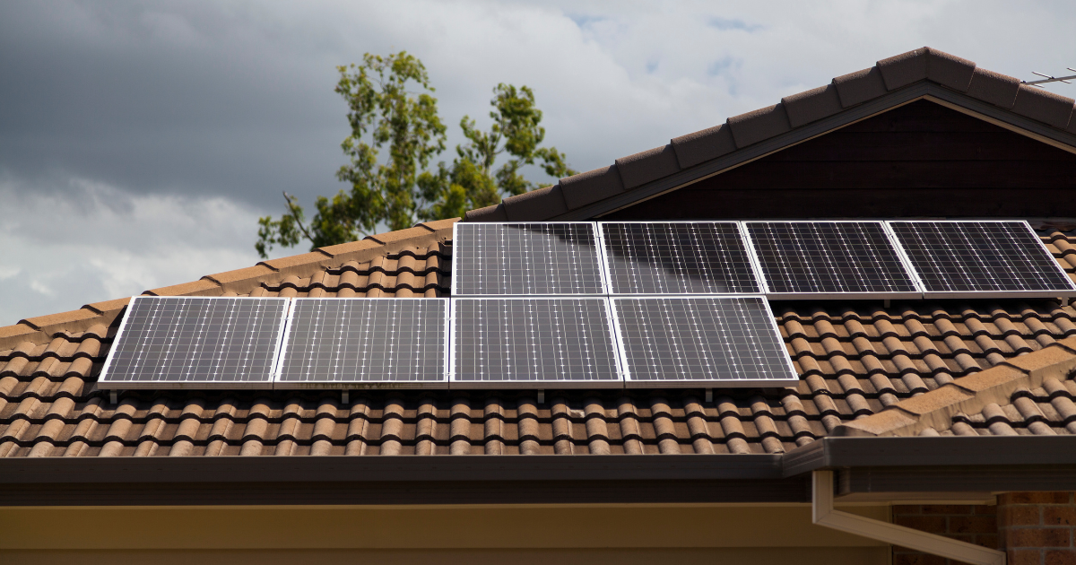  Telhado de casa com painéis solares instalados sobre telhas cerâmicas marrons. O fundo tem céu nublado e árvores, destacando a energia sustentável.