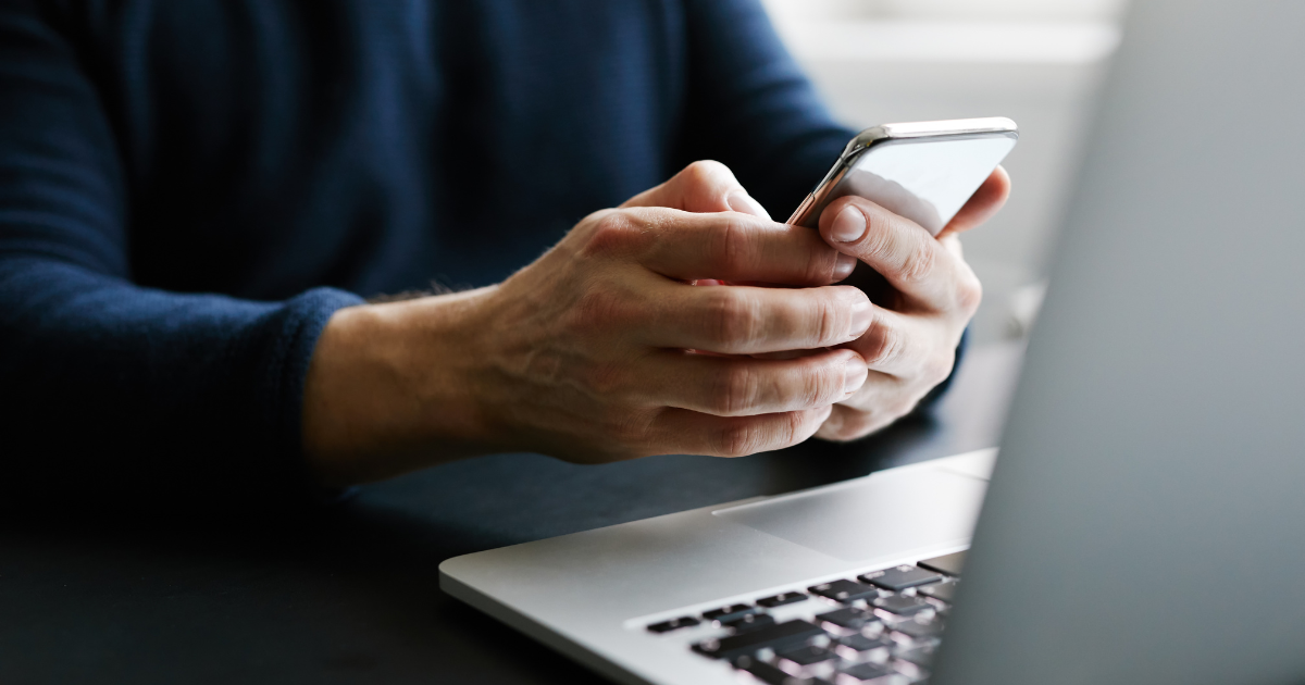 Pessoa segurando um smartphone próximo a um notebook, sugerindo uso de tecnologia ou trabalho remoto.