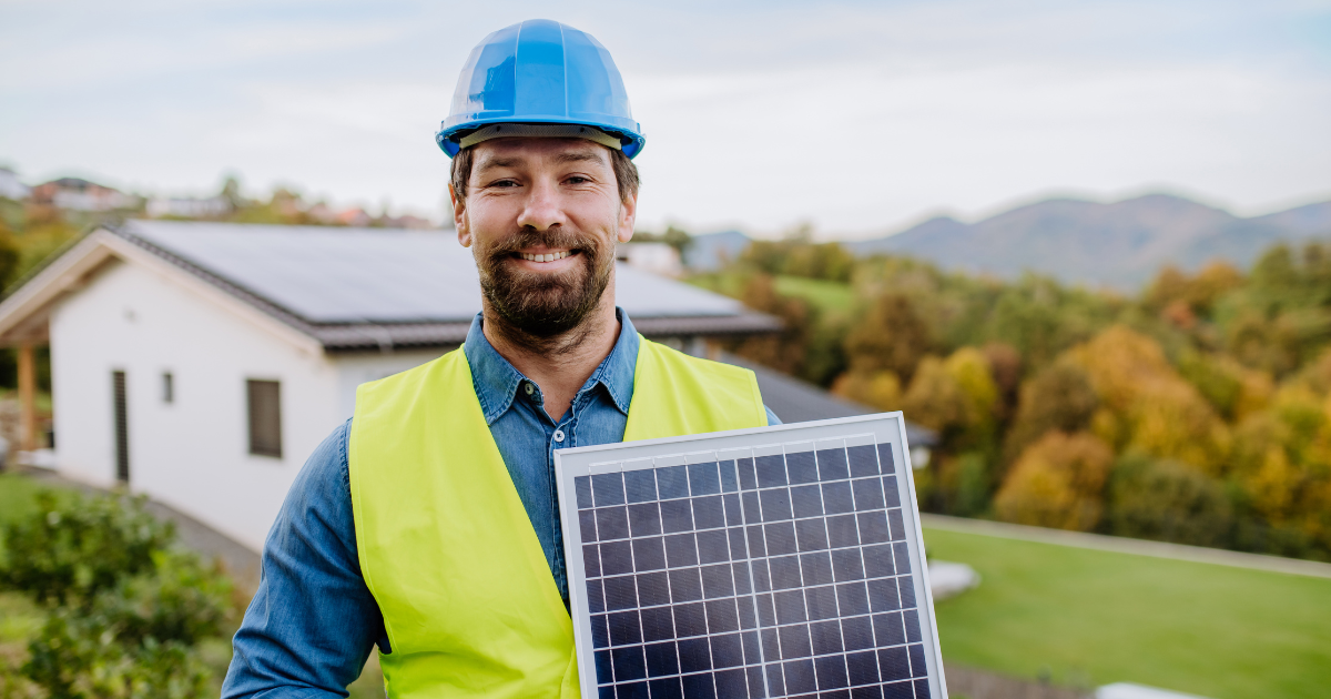 Empreendedor solar com capacete azul e colete amarelo segura um painel solar, com uma casa e montanhas ao fundo.
