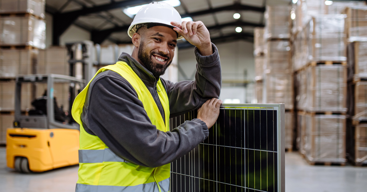 Empreendedor solar com capacete branco e colete amarelo apoia-se em um painel solar dentro de um armazém com caixas empilhadas.