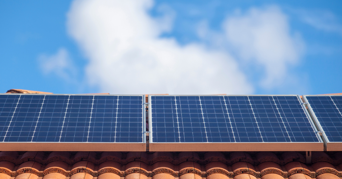 Painéis solares sobre telhas cerâmicas sob um céu azul com nuvens. Representa o uso de energia limpa e sustentável.