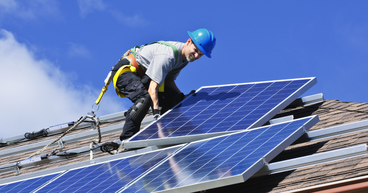 Empreendedor solar com equipamentos de segurança instala painéis solares no telhado sob céu azul. A imagem representa energia solar e trabalho especializado.