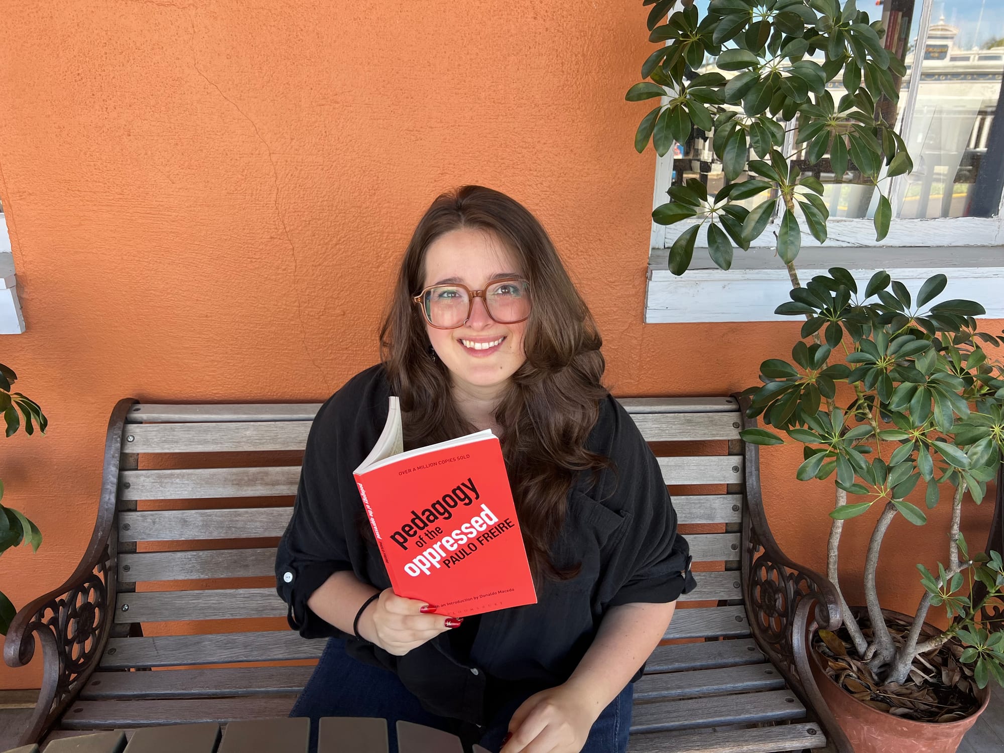 Anna Platt sits on a bench while holding Paulo Friere's book, Pedagogy of the Oppressed. She is wearing a black button-down shirt and large red glasses.
