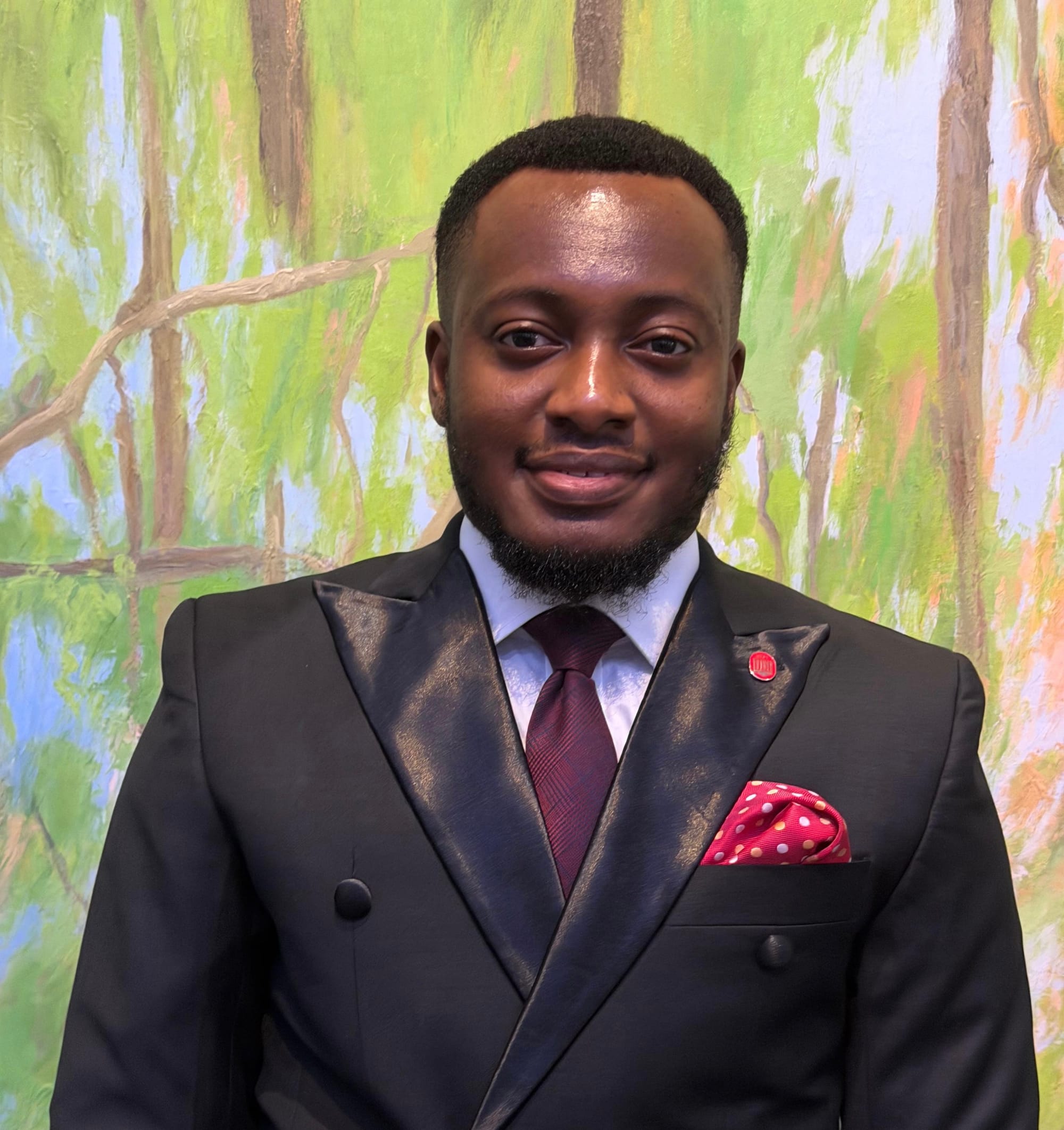 Godwin Egbe appears in front of a mural. He is wearing a suit with a burgundy tie and red polka dotted pocket hankerchief.