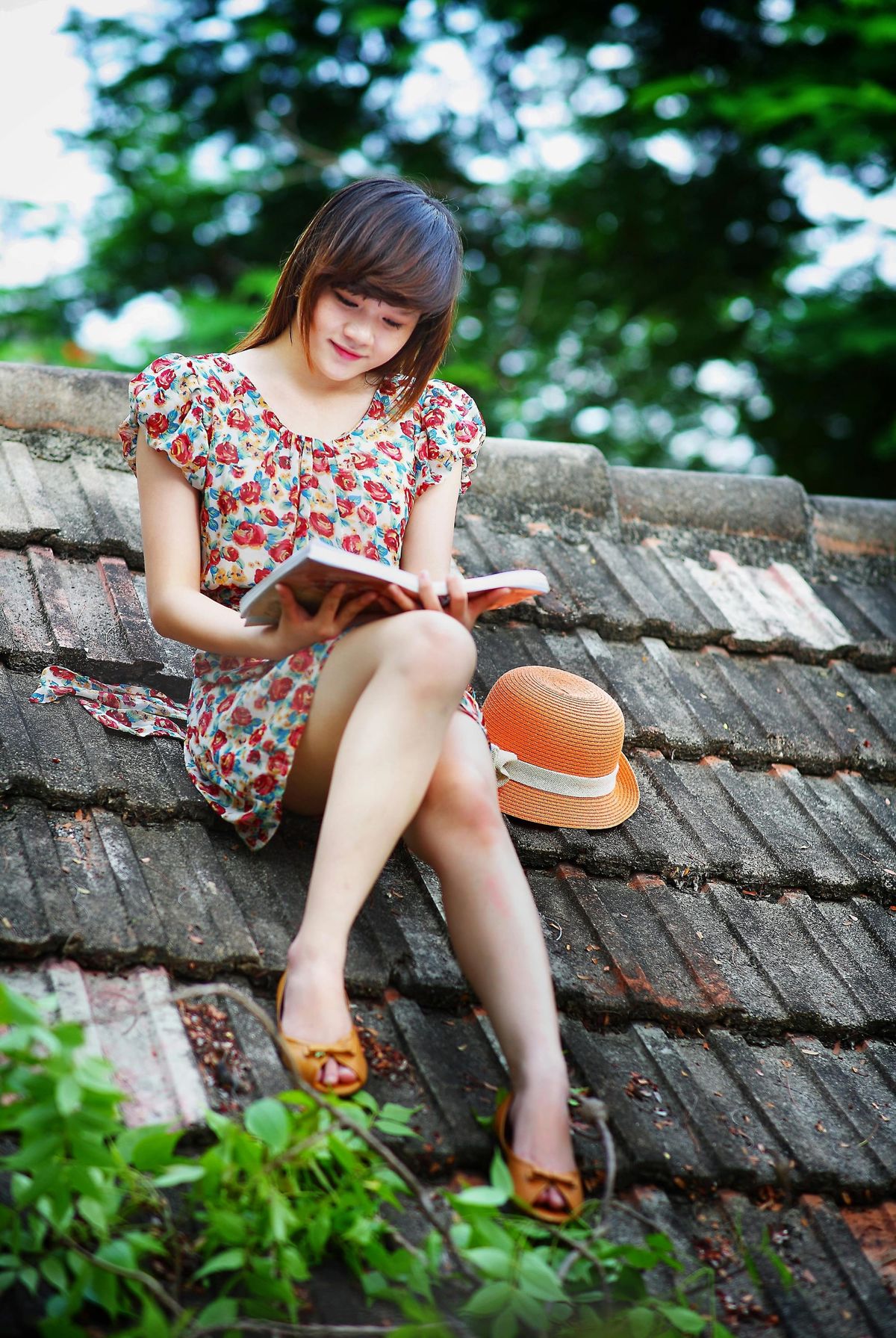 A woman wearing a white, green, and red floral cap-sleeved dress is reading a book on black roof near green trees.