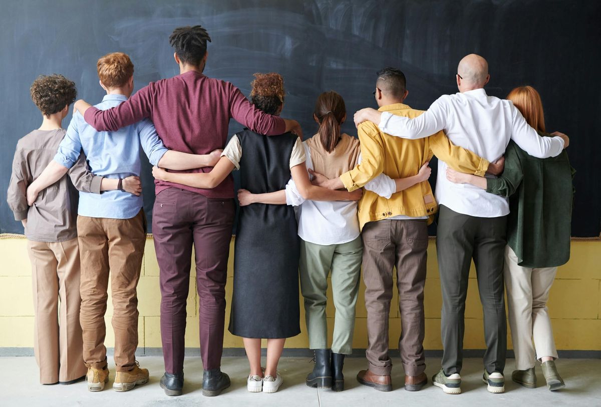 a group of people of various heights and apparent ethnicities is shown standing while facing a blalckboard. We see them from behind, and their arms are linked around one another's shoulder