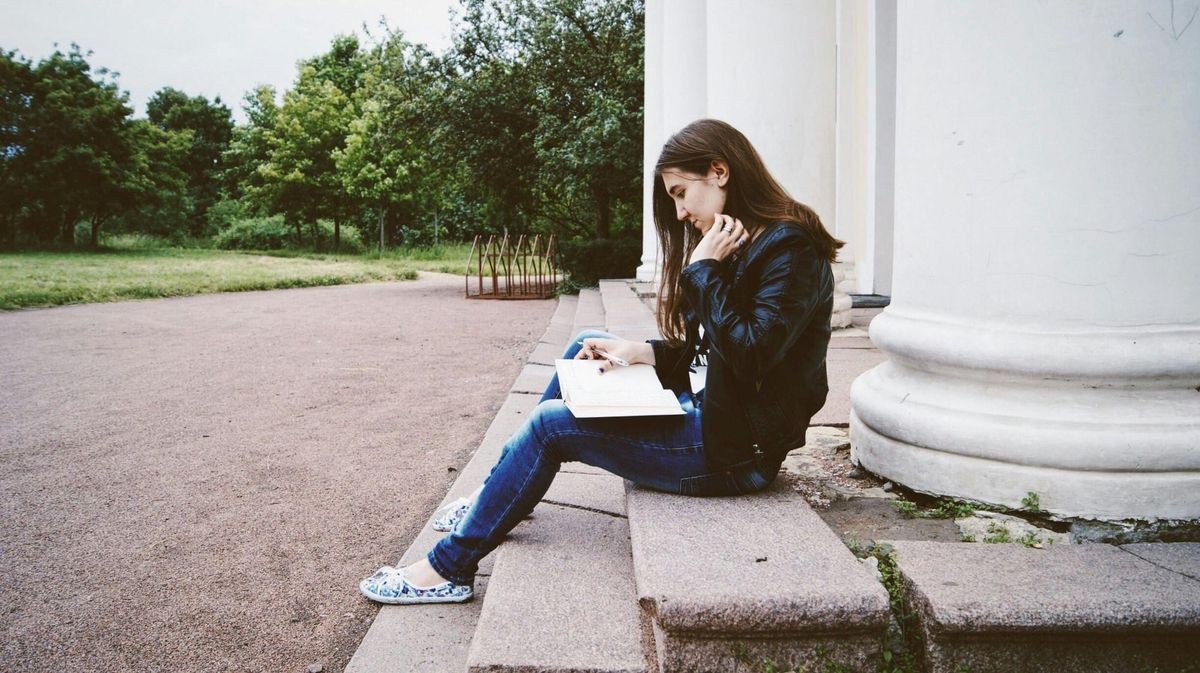 A white woman with long brown hair, wearing a leather jacket, jeans, and canvas shoes, sits on a step in front of a white pillar while reading, pen in hand.