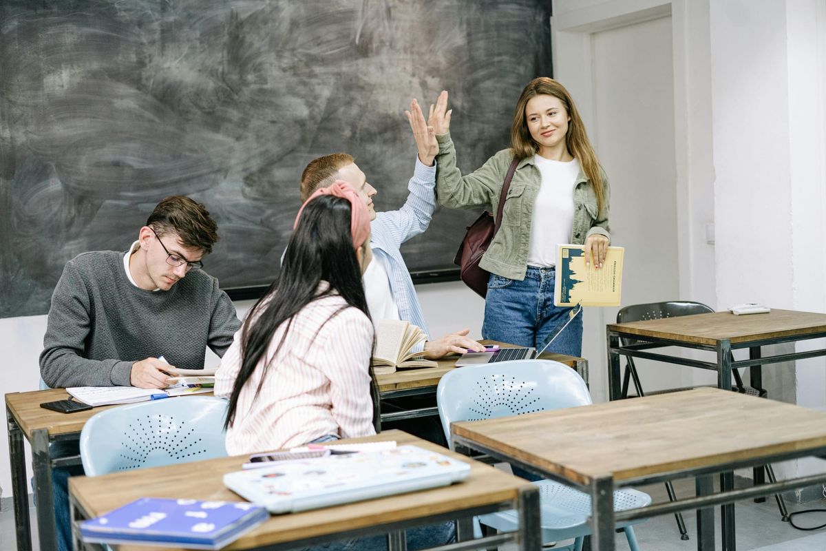 Four college students are in a classroom with a chalkboard in the background and single desks + chairs arranged in rows. Two of the people are giving each other a high five.