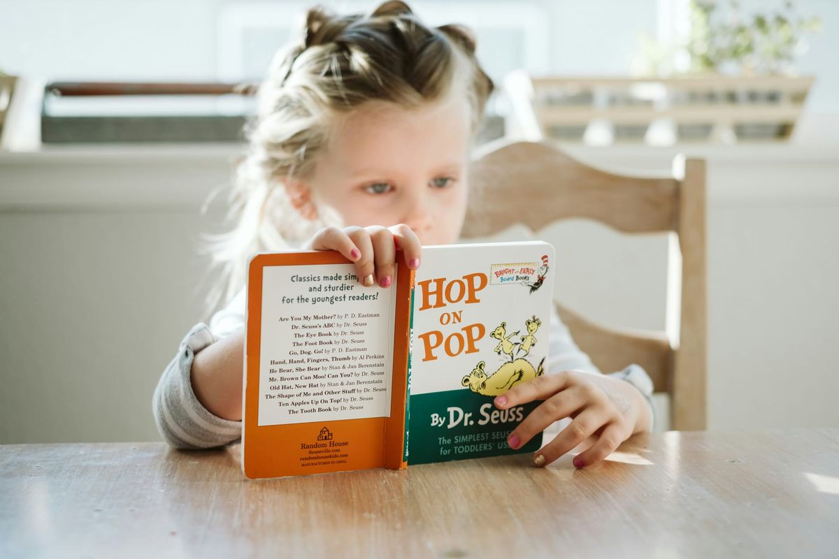 A girl sits at a table, reading "Hop on Pop" by Dr. Seuss.