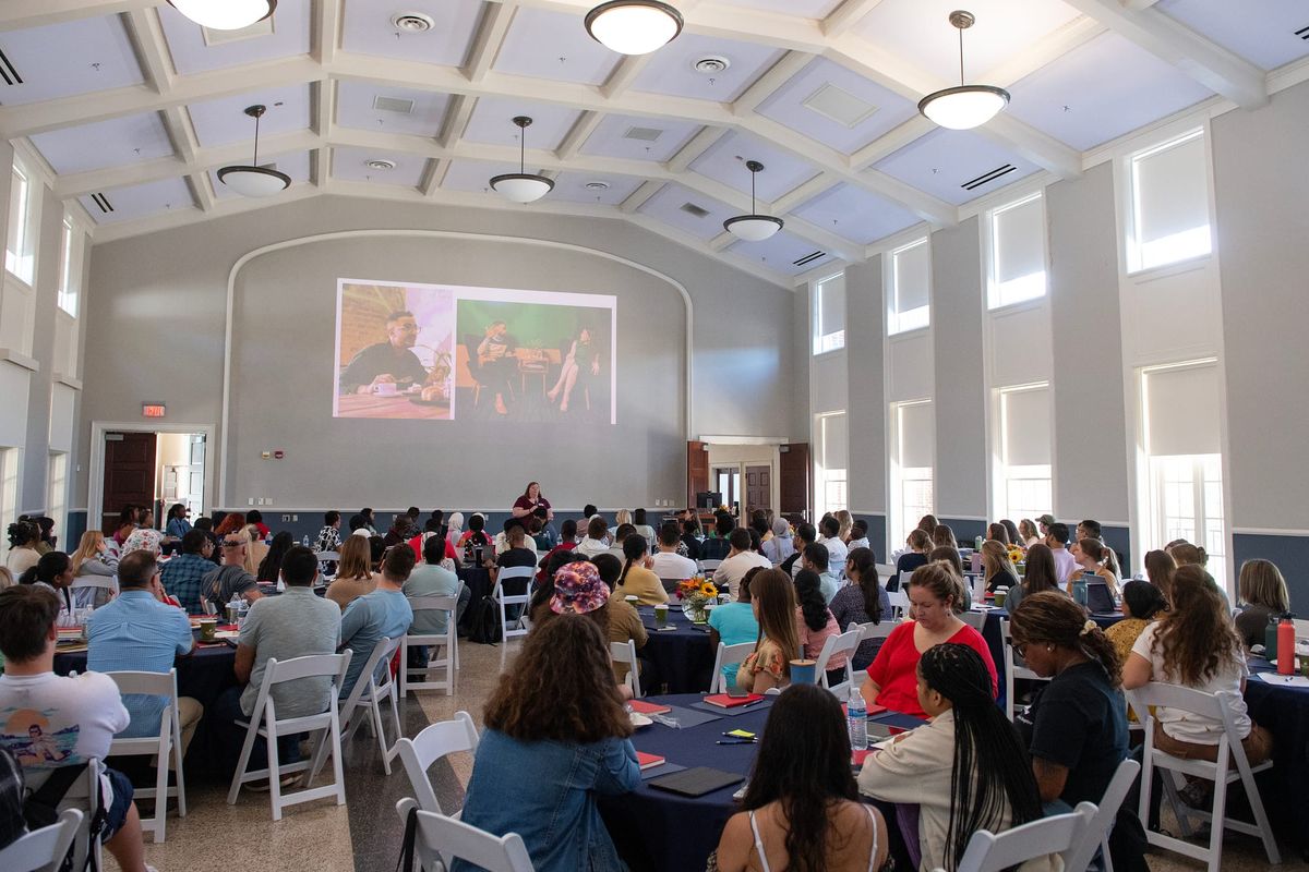 A large number of students attend a workshop in a ballroom. They are sitting at round tables with blue tablecloths.