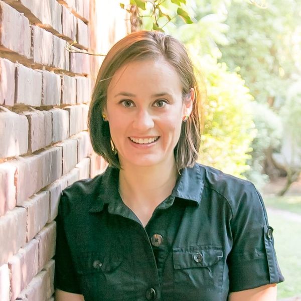Summer Hill-Vinson is seen standing near a brick wall in a darkly colored button-down short-sleeved shirt, in front of a green outdoor space.