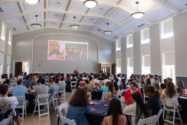 A large number of students attend a workshop in a ballroom. They are sitting at round tables with blue tablecloths.