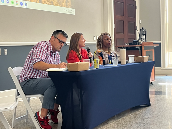 Three people at a table draped in blue: Joel Amidon (a white man w/plaid shirt), Nicole Roberson (a white woman w/red shirt), and Candies Winfun-Cook (a Black woman w/multicolor shirt).