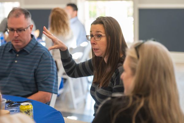 A woman in the center of the picture has a hand up as she makes a point while discussing with two others. In the background are other tables of faculty discussing.