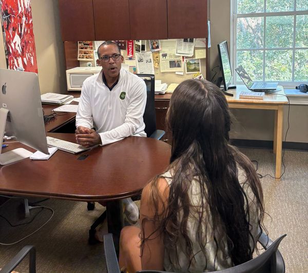 Marvin King, a Black instructor in a white quarter-zip shirt, sits at his desk. A student in a chair facing him is shown. She has long dark hair.