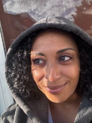 Woman with curly dark hair wearing a gray hooded sweatshirt, looking off to the side with a slight smile and natural lighting.