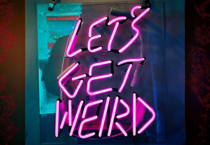 Bright pink neon ‘LET’S GET WEIRD’ sign mounted on dark wall.