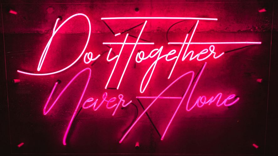 Neon sign reading "Do it together, Never Alone" in pink and white script against a dark concrete wall.