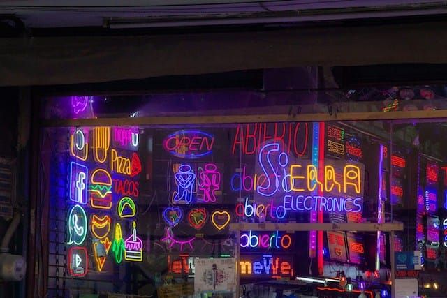 Storefront window filled with multiple colorful neon signs in various languages creating a vibrant display of different colored lights and text.