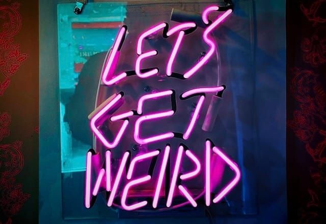 Bright pink neon ‘LET’S GET WEIRD’ sign mounted on dark wall.