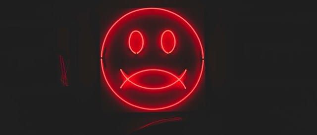 A red neon face emoji glowing against a dark background. The face has two oval eyes and a both an upward and downward curved mouth.