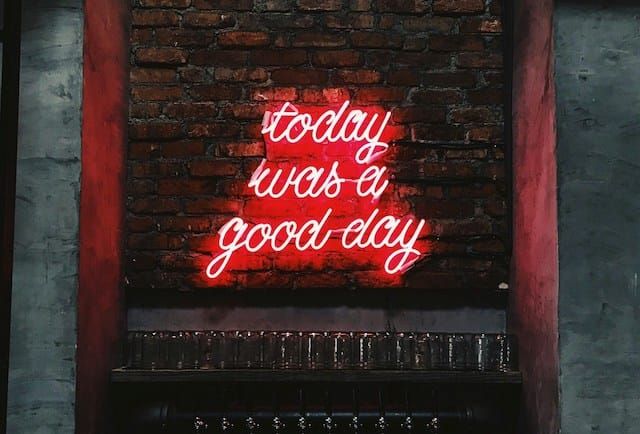Neon sign on a brick wall that reads "today was a good day" in cursive, glowing red. The sign is above a shelf lined with glassware in a dimly lit space.
