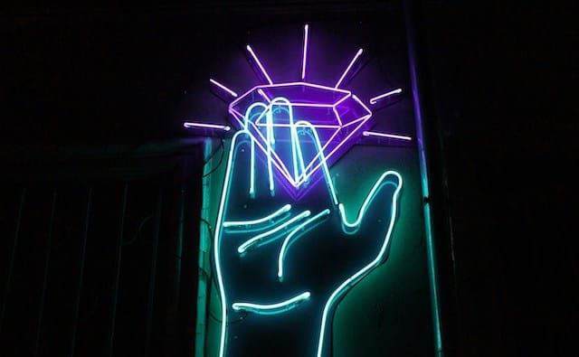 Neon artwork of a teal hand reaching for a purple diamond, glowing against a dark background.