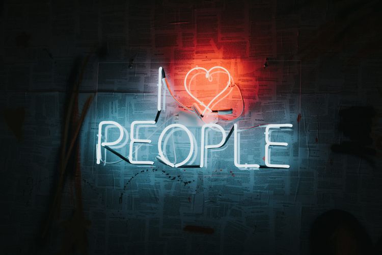 Neon sign reading "I ♥ PEOPLE" in white and red against a wall covered with newspaper clippings