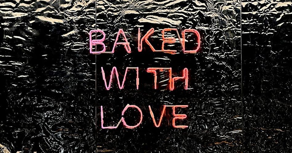 Neon sign reading "Baked With Love" in pink and red letters against a dark reflective background