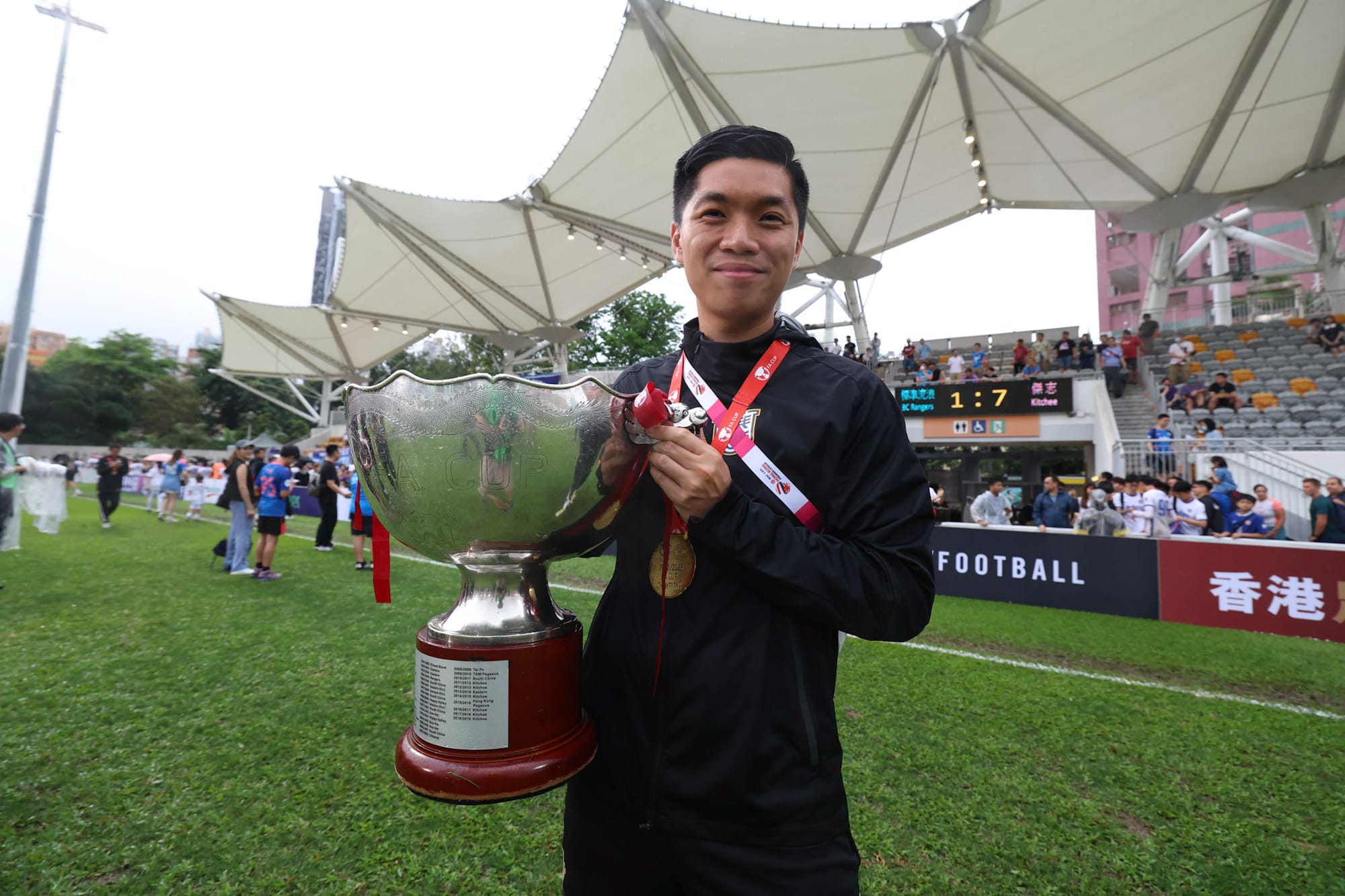 Norris Ng holding the silver Hong Kong FA Cup trophy and wearing a winner's medal in a black Kitchee FC training jacket, with the final scoreboard reading BC Rangers 1 – 7 Kitchee in the background.